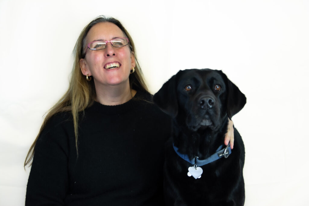Zwart-wit foto van Debby die samen met zwarte labrador Tarka poseert voor een witte achtergrond. Debby zit links in de foto, draagt een zwarte trui, een bril en glimlacht vrolijk met gesloten ogen. Ze lijkt in een oprechte lach te zijn vastgelegd. Haar linkerhand houdt Tarka op een liefdevolle manier vast. Ze heeft haar linkerhand onder de kin van Tarka, wat bijdraagt aan een gevoel van nabijheid en kameraadschap tussen de twee. Tarka zit rechts en kijkt recht in de camera met een alerte en rustige uitdrukking. Haar vacht is egaal en glanzend. Tarka draagt een halsband. De foto ademt een sfeer van warmte en vriendschap uit.