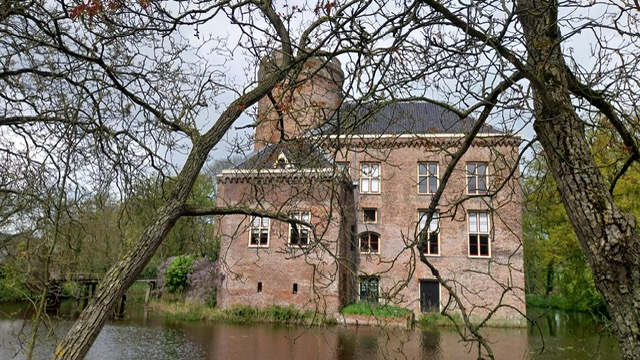 Foto van Kasteel Loenersloot. Het is een oud, schilderachtig kasteel met een grote ronde toren. Het kasteel is gebouwd van rode bakstenen en heeft meerdere ramen met witte kozijnen. Het dak van de toren lijkt bedekt met donkere dakpannen. Het kasteel is omgeven door een gracht met helder water en weelderige groene bomen, waarvan sommige hun takken over het water uitstrekken. Het lijkt een rustige, historische locatie.