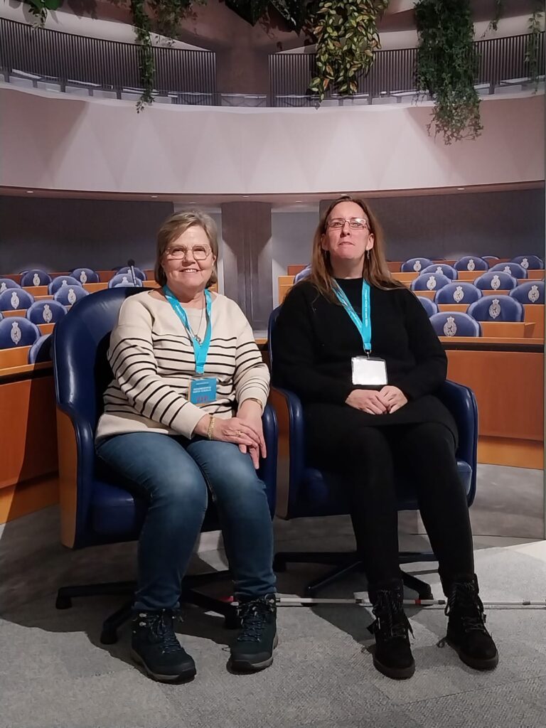 Foto van Debby en haar vriendin die op donkerblauwe stoelen met een hoge rugleuning zitten bij de selfiespot in de Tweede Kamer. Debby’s vriendin (links) heeft kort haar, draagt een bril, een gestreepte trui en een donkere broek. Ze glimlacht naar de camera. Debby (rechts) heeft lang haar, draagt een zwarte top en een zwarte broek, en kijkt serieus voor zich uit. Door de achtergrond lijkt het of ze als Kamerleden in de Tweede Kamer zitten. De stoelen lijken comfortabel en zijn opgesteld in rijen, zoals je vaak ziet in een conferentie- of vergaderzaal.
