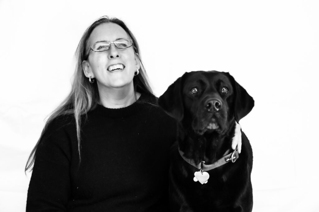 Zwart-wit foto van Debby en zwarte Labrador Tarka. Ze zitten voor een witte achtergrond. Debby kijkt naar Tarka en lacht. Ze heeft een bril op en draagt een zwarte trui en legging. Tarka, die een halsband met een hartvormig naamplaatje om heeft, kijkt recht in de camera met een alerte blik. De foto is in zwart-wit, wat voor een klassiek en tijdloos effect zorgt.