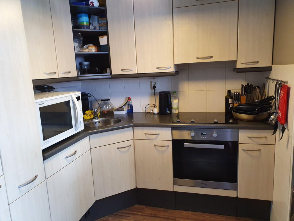Foto van de keuken van Debby. Het aanrechtblad is grijs en de keukenkastjes zijn geelwit. Op het aanrecht staan een magnetron en een waterkoker. Er is een zwarte inductiekookplaat te zien. Onder de kookplaat zit een inbouwoven.