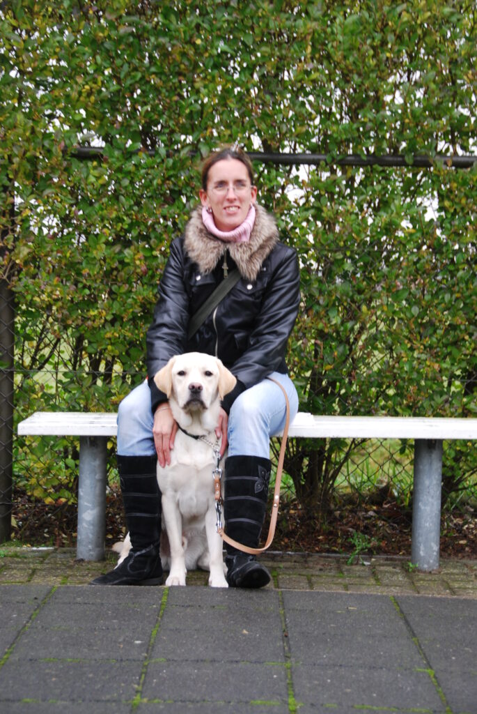 Foto van Debby, die tijdens de schoolinstructie in december 2009, op een bankje zit bij KNGF Geleidehonden met tussen haar benen een zittende zeer blonde labrador, Umbar.