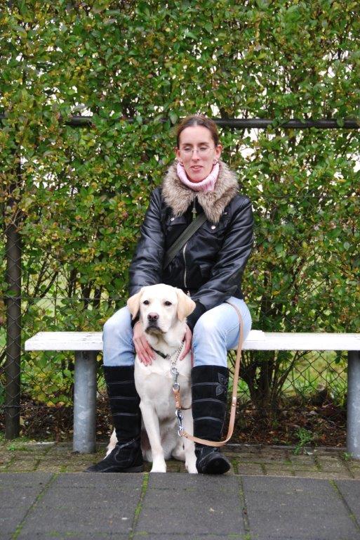Foto van Debby, die tijdens de schoolinstructie in december 2009, op een bankje zit bij KNGF Geleidehonden met tussen haar benen een zittende zeer blonde labrador, Umbar.