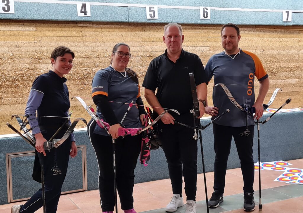 Foto van Angela en haar handboogteam. Ze staan met z’n vieren voor een wand met schietdoelen. Alle vier hebben ze hun boog vast en om hun middel dragen ze een pijlentas, waar pijlen in zitten. Van links naar rechts zie je: Lorenza, Angela, Rob en Thijs.