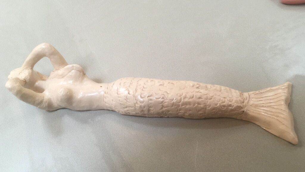 Foto van Debby’s eerste kleiwerkstuk. Het is een beeld van een zeemeermin, die languit op haar rug ligt en haar handen voor haar gezicht houdt, waardoor haar ellebogen omhoogsteken. De zeemeermin heeft lang haar, waarvan een haarlok over haar borst ligt. De zeemeermin is na het afbakken in de oven afgewerkt met caparol, waardoor haar details extra duidelijk zijn.