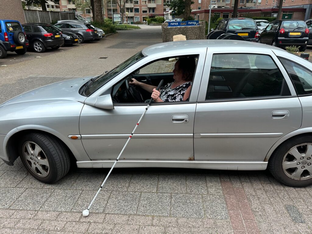 Foto van een zilvergrijze auto op een parkeerplaats met Miriam Taylor achter het stuur. Uit het open raam steekt haar taststok, waarmee ze over de grond voelt.