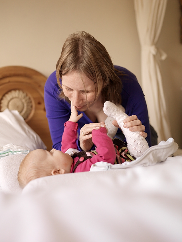 Foto van Elian die over een baby heen buigt die op een bed ligt. Elian draagt een blauwe trui en heeft bruin haar. Ze houdt haar hand onder haar kin en kijkt bedachtzaam naar de baby. De baby ligt op haar rug en houdt een witte sok vast. Ze kijkt naar Elian en lijkt een interactie met haar te hebben. Het beddengoed is wit en er is een deel van een houten hoofdeinde en een wit gordijn op de achtergrond zichtbaar