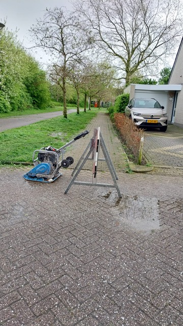 Foto van een straat met een stoep en een rij bomen. De stoep wordt geblokkeerd door een bouwhek. Op de voorgrond ligt een blauwe trilplaat op zijn kant op een bestrating van grote vierkante stenen. Er is een driepoot wegwijzerafzetting met een verkeersbord direct naast de trilplaat. Achter de verkeersafzetting zie je een waterplas op de bestrating. De omgeving is groen met bomen in blad en wat begroeiing langs de randen van de weg.