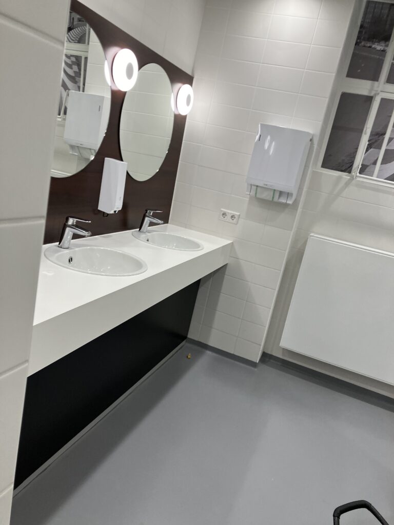 Foto van een nette, moderne, openbare toiletruimte. De ruimte heeft een witte tegelmuur met grote rechthoekige tegels. In het midden van de afbeelding is een wastafelmeubel met twee ronde wastafels ingebouwd in een wit werkblad zichtbaar. Boven elke wastafel hangt een ronde spiegel bevestigd aan een houten achterwand, met boven elke spiegel een ronde lamp die een helder licht uitstraalt. Tussen de wastafels hangt een automatische zeepdispenser aan de muur. Rechts van de wastafels hangt een witte handdoeken- of papieren-handdoekdispenser aan de wand. Boven de dispenser bevindt zich een raam met een metalen kozijn. Onderaan de muur is een radiator zichtbaar. De vloer is grijs met een effen afwerking. Naast de handdoekautomaat is een doorgang naar de toilethokjes.