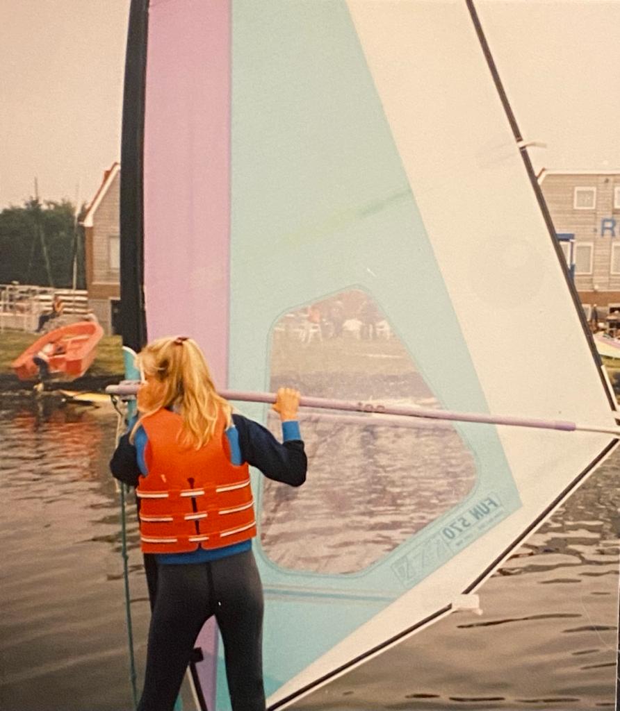 Foto van Elian als tiener die aan het windsurfen is. Ze heeft blond haar en draagt een reddingsvest in de kleuren oranje en blauw, en een zwarte wetsuit. Ze staat met haar rug naar de camera en houdt een windsurfzeil vast dat roze en lichtblauw van kleur is. Op de achtergrond is water te zien met een andere windsurfer en enkele huizen aan de overkant van het water. Het lijkt een bewolkte dag te zijn.