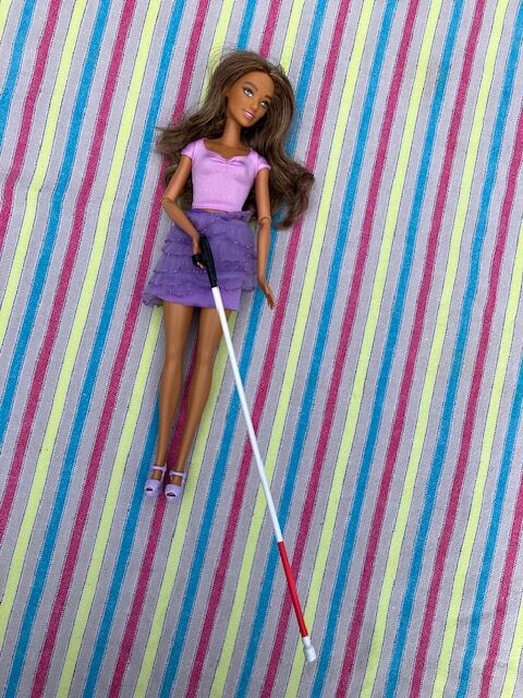 Foto van een Barbie die op een gestreept oppervlak ligt. De pop heeft lang, golvend donkerblond haar en draagt een lichtpaars shirtje met korte mouwen en een ronde hals. Ze heeft een paarse rok aan die uit meerdere laagjes bestaat met een ruche-achtige afwerking. Aan haar voeten draagt ze lichtpaarse schoenen met hoge hakken. De pop houdt een blindenstok vast en ze heeft een zonnebril boven op haar hoofd. De achtergrond bestaat uit een kleurrijk gestreept patroon met verticale strepen in de kleuren roze, blauw, geel en wit.