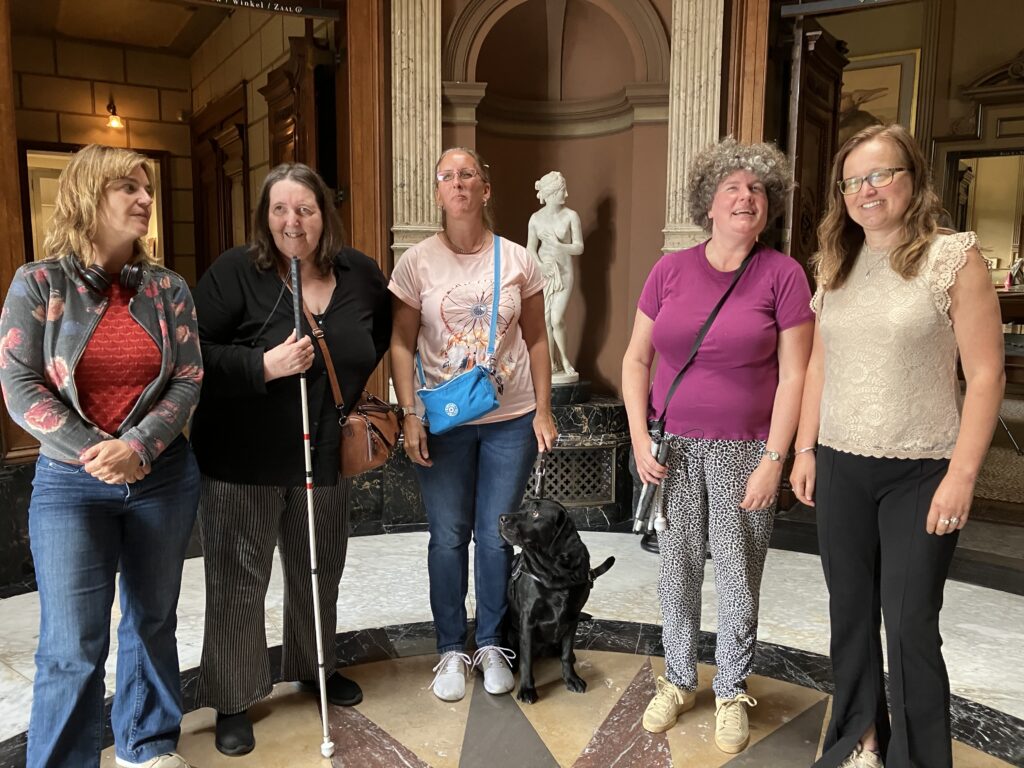 Foto van Debby en haar geleidehond Tarka, die met een groepje vrouwen in de ontvangsthal van het Teylers Museum staat. Het is een elegante, klassiek ingerichte ruimte met een marmeren vloer met een geometrisch patroon. Achter hen zijn een klassiek marmeren standbeeld van een naakte vrouw en zuilen te zien. De vrouwen zijn casual gekleed en sommige van hen hebben accessoires zoals een handtas of witte stok.
