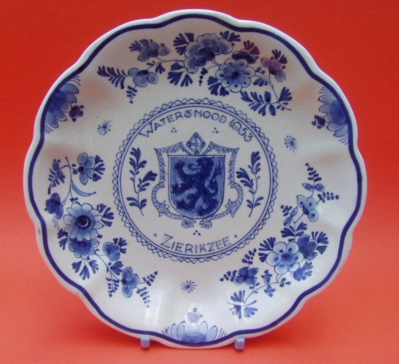 Foto van een decoratief bord in Delfts blauwe stijl. Het bord heeft een golvende rand en het heeft een witte achtergrond met daarop blauwe decoraties. In het midden van het bord is een cirkelvormig embleem met daarin een wapenschild met een leeuw te zien. Boven het wapenschild staat de tekst ‘WATERSNOOD 1953’ en eronder staat ‘ZIERIKZEE’. Rondom het centrale embleem is het bord versierd met een traditioneel Delfts blauw bloemenpatroon. Dit patroon bestaat uit verschillende bloemen, bladeren en ranken in verschillende tinten blauw. Het bord staat rechtop op een felrode achtergrond. Dit bord is een herdenkingsstuk voor de watersnoodramp van 1953.