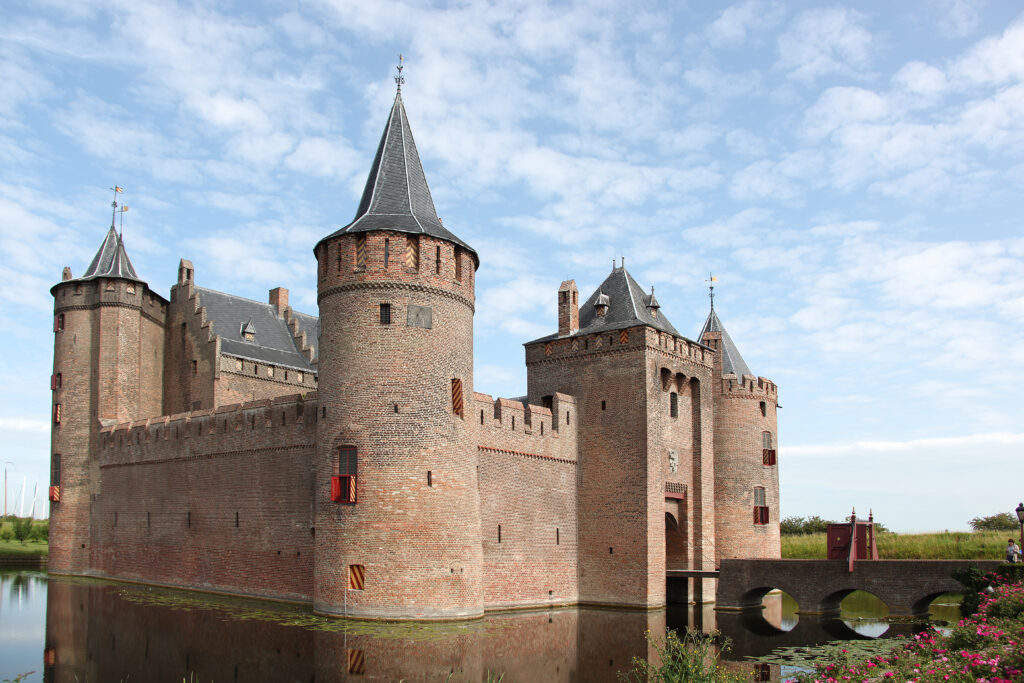 Foto van de buitenkant van het Muiderslot. Het middeleeuwse kasteel ligt aan het water en is omringd door een slotgracht. Het is gebouwd van rode bakstenen en heeft ronde torens met spitse, donkergrijze daken. De torens hebben kleine vensters met decoratieve rode luiken en boven op de torens zitten kantelen. De burcht is toegankelijk via een stenen boogbrug over de gracht. De weerspiegeling van het kasteel in het water van de gracht creëert een schilderachtig effect. De lucht op de foto is helderblauw met witte wolken. Aan de oever van de gracht zijn roze bloemen te zien.