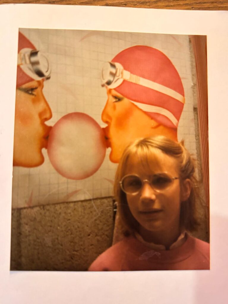 Foto van een ongeveer 10-jarige Elian. Ze heeft blond haar en draagt een roze trui en een bril. Boven haar hangt een poster aan de muur met daarop twee naar elkaar toegebogen meisjesgezichten met elk een roze badmuts en een zwembril op. Samen blazen ze één grote roze kauwgombel.