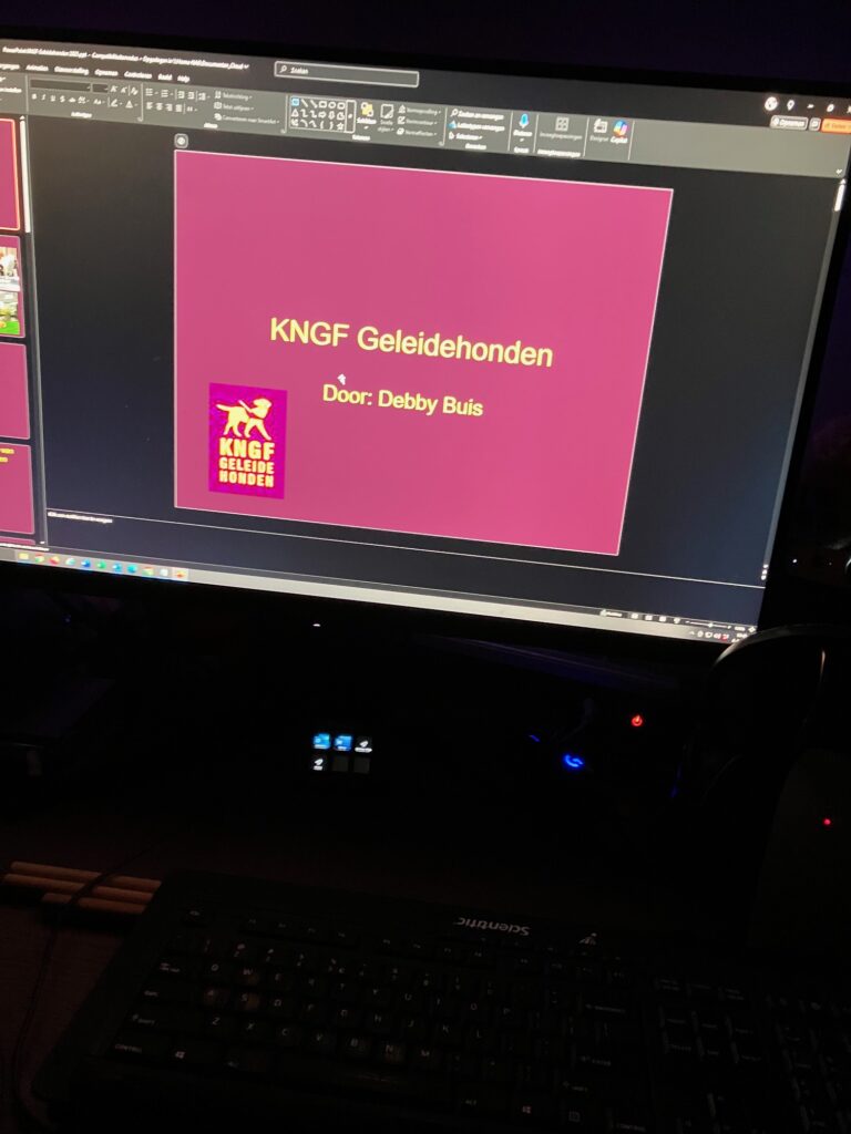 Foto van een computerscherm met een presentatie die geopend is in Microsoft PowerPoint. De dia heeft een roze/paarse achtergrond en bevat het logo van ‘KNGF Geleidehonden’. Het KNGF-logo toont een gestileerde afbeelding van een hond in geel/goud op een paarse achtergrond. Onder de titel ‘KNGF Geleidehonden’ staat ‘Door: Debby Buis’. Het scherm toont ook de PowerPoint werkbalk bovenaan met verschillende bewerkingstools. Aan de zijkant zijn miniaturen van andere dia's te zien.