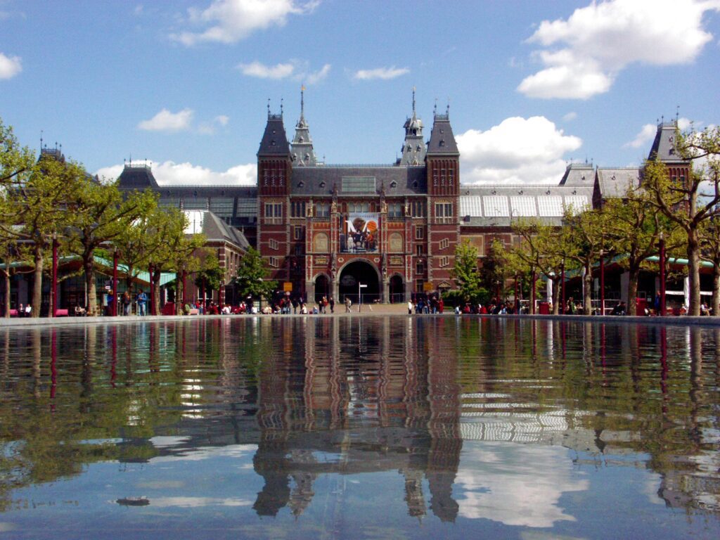 Foto van het Rijksmuseum in Amsterdam. De foto toont de voorkant van het majestueuze bakstenen gebouw in neogotische stijl, met zijn karakteristieke rode bakstenen façade en twee prominente torens. Op de voorgrond is een ondiepe waterpartij te zien die zich voor het museum bevindt. Het water creëert een perfecte weerspiegeling van het gebouw en de wolken in de lucht, wat een prachtig symmetrisch effect geeft. De foto is genomen op een mooie dag met een helderblauwe lucht en enkele witte wolken. Aan weerszijden van het gebouw staan bomen die net in blad lijken te staan, wat suggereert dat de foto in de lente is genomen. Er zijn verschillende mensen zichtbaar op het plein. Het gebouw heeft een imposante centrale toegangspoort met een boog, geflankeerd door twee hoge torens met spitse daken. De architectuur is zeer gedetailleerd met vele ornamenten, kantelen en decoratieve elementen die typerend zijn voor de 19e-eeuwse bouwstijl.