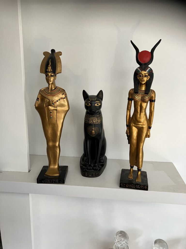 Foto van drie Egyptische decoratieve beelden die op een witte plank staan. Links staat een gouden beeld van Osiris in zijn kenmerkende mummievorm met een kroon op zijn hoofd en een kromstaf en vlegel gekruist voor zijn borst. In het midden staat een zwart beeld van een Egyptische kat (Bastet), rechtop zittend met versieringen en hiërogliefen op de borst. Rechts staat een gouden beeld van de godin Isis, gekenmerkt door haar slanke figuur en de rode zonneschijf met hoorns op haar hoofd. Alle drie de beelden staan op zwarte voetstukken met hiërogliefen erop. Ze staan tegen een witte achtergrond op een plank bij Debby thuis.