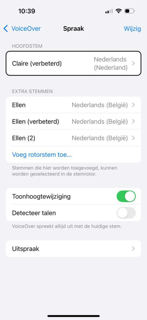 Screen-shot van de instellingenpagina van VoiceOver tabblad ‘Spraak’ op een iPhone. De hoofdsectie heet ‘Hoofdstem’ en daar is ‘Claire (verbeterd)’ geselecteerd met als taal ‘Nederlands (Nederland)’. Daaronder staat ‘Extra stemmen’ met drie opties: Ellen - Nederlands (België), Ellen (verbeterd) - Nederlands (België) en Ellen (2) - Nederlands (België). Daaronder staat een blauwe link: ‘Voeg rotorstem toe...’ Verder naar beneden zijn er twee schakelaars: ‘Toonhoogtewijziging’ (staat aan, groen) en ‘Detecteer talen’ (staat uit, grijs). Onderaan staat de optie ‘Uitspraak’. Er is uitlegtekst: ‘Stemmen die hier worden toegevoegd, kunnen worden geselecteerd in de stemrotor.’ en ‘VoiceOver spreekt altijd uit met de huidige stem.’