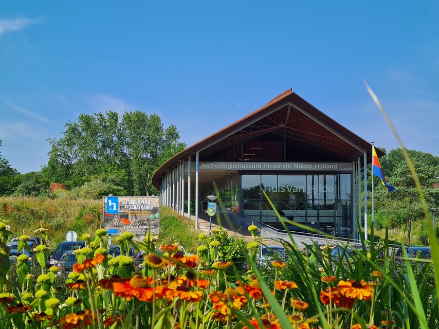 Foto van de buitenkant van Archeologiemuseum Huis van Hilde in Castricum. Het gebouw heeft een moderne architectuur met een opvallend schuin dak en grote glazen raampartijen aan de voorkant. De entree is duidelijk zichtbaar met de naam van het museum op de gevel. Op de voorgrond zijn oranje bloemen te zien die in volle bloei staan. De omgeving van het museum is zeer groen met bomen en natuurlijke vegetatie op de achtergrond. Er is ook een informatiebord/reclamebord zichtbaar naast het gebouw. Er is ook een vlag zichtbaar bij het gebouw. Het parkeerterrein is gedeeltelijk zichtbaar en het hele tafereel straalt een verzorgde en professionele uitstraling uit. Het is een zonnige dag met een helderblauwe lucht.
