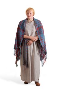 Foto van de reconstructie van Hilde in het Archeologiemuseum Huis van Hilde. Het is een vrouw met rossig haar in een knot, die historische kledij draagt. Ze draagt een lange, eenvoudige beige of grijze tuniek die tot op de enkels valt. Om haar schouders ligt een opvallend geruite rood-blauwe mantel, met franjes aan de randen. De mantel heeft een traditioneel Schots tartanpatroon. Aan haar voeten draagt ze eenvoudige, bruine, platte, leren schoenen. Om haar taille heeft ze een geweven of gevlochten ceintuur in dezelfde kleuren als de mantel. Ze draagt een ketting. De reconstructie staat voor een witte achtergrond.
