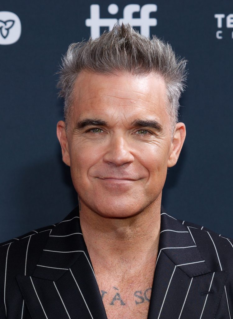 Foto van Robbie Williams op de rode loper van het Toronto International Film Festival in 2024. Hij is een man van middelbare leeftijd met opvallend grijs/zilverkleurig haar dat stijlvol omhoog is gestyled. Hij draagt een zwart pak met dunne witte krijtstrepen en heeft het jasje open, waaronder een deel van zijn ontblote borst met wat tatoeages zichtbaar is. Hij heeft een warme, zelfverzekerde glimlach en heldere groene ogen. De achtergrond is donkerblauw en er zijn wat logo's vaag zichtbaar.