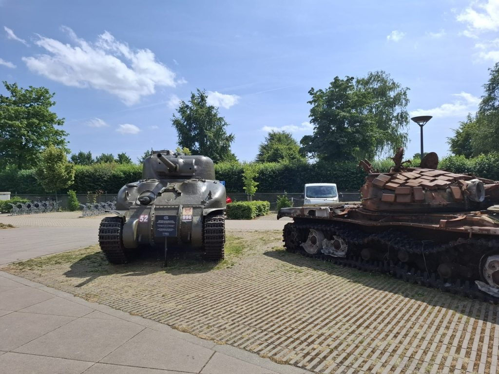 Foto van twee tanks die buiten staan tentoongesteld bij het Vrijheidsmuseum in Groesbeek. Links staat een goed onderhouden olijfgroene Amerikaanse Sherman tank uit WO II, gemaakt van metaal met verschillende details zoals nummerplaatjes met de nummers 52 en 996 erop. Rechts staat een roestbruine, vernielde Russische tank uit Oekraïne, die er roestig uitziet. Op de achtergrond staat een omheinde groene heg met bomen en struiken, en enkele fietsrekken. De hemel is blauw met een paar wolken en de zon schijnt helder. Er is ook een klein wit voertuig tussen de tanks en de heg te zien.