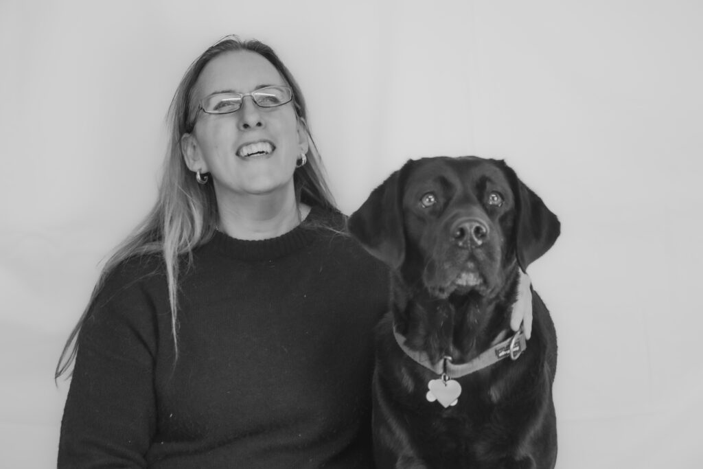 Zwart-wit foto van Debby en haar blindengeleidehond Tarka. Debby lacht met haar ogen dicht en draagt een bril en een lange, zwarte trui. Tarka zit naast haar. Tarka is een zwarte labrador retriever en draagt een halsband met een hondenpenning. Ze kijken beide recht in de camera en achter hen is een egale, lichte achtergrond. Ze lijken opgewekt en gelukkig samen.