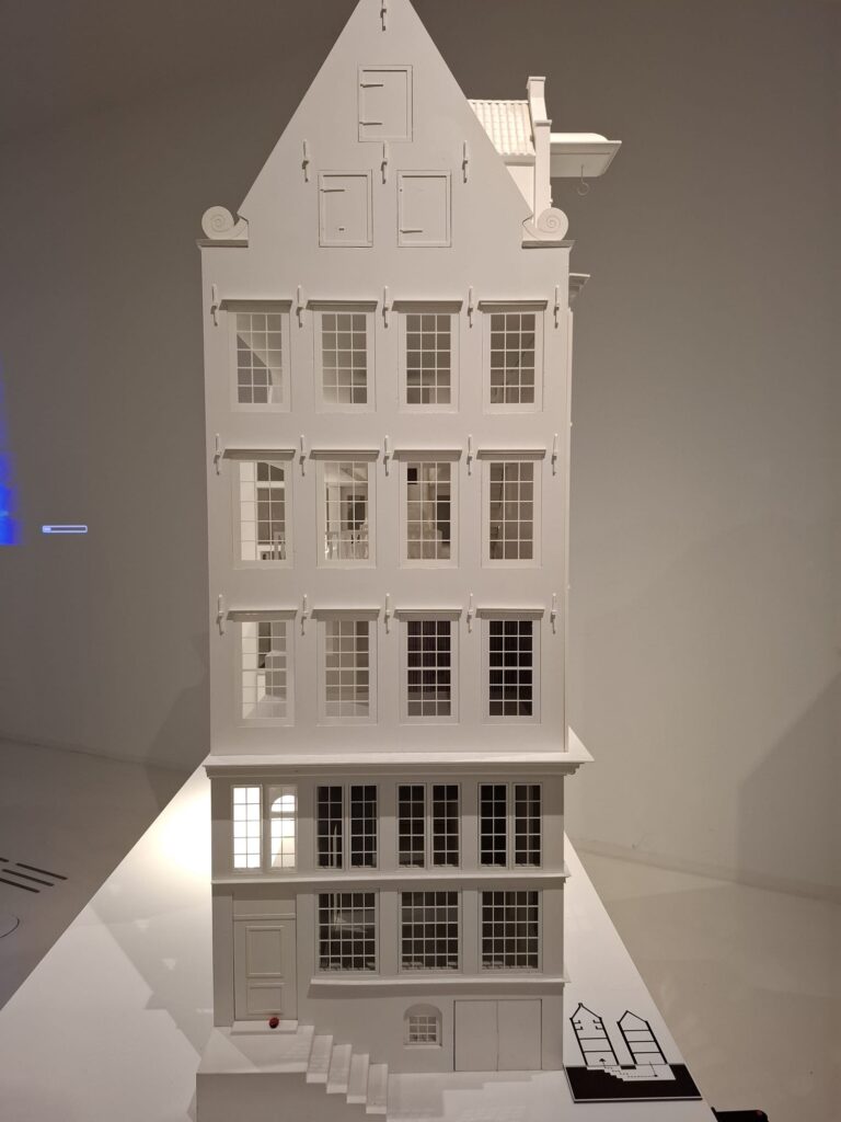Foto van een gedetailleerde, witte maquette van de voorgevel van Museum Ons’ Lieve Heer op Solder. Het is een traditioneel Amsterdams grachtenhuis. Het is smal en hoog en heeft een puntdak, karakteristieke ramen met kleine ruitjes en luiken boven de ramen. De gevel is versierd met een puntgevel, voorzien van drie kleine ramen of luiken en sierlijke krullen (voluten) aan weerszijden. Daaronder bevinden zich drie verdiepingen met elk vier grote ramen met roedeverdeling. Op de begane grond zijn grote raampartijen te zien en een trapje dat leidt naar de voordeur. Onder de voordeur ligt een souterrain met een toegangsdeur en kleinere ramen.
