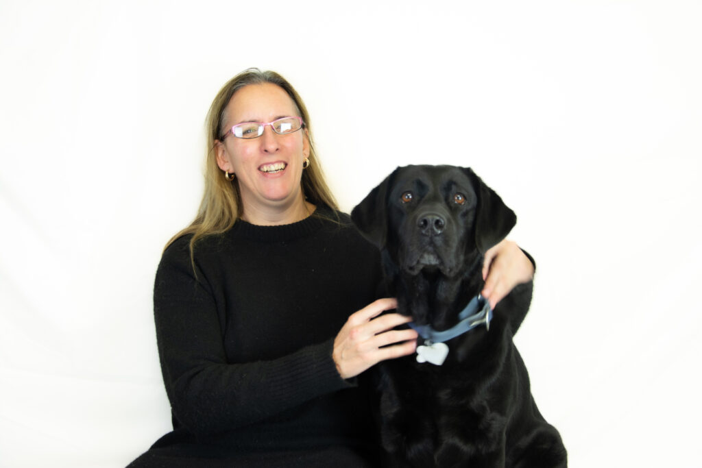 Foto van Debby en haar blindengeleidehond Tarka. Debby draagt een zwarte trui en een bril en ze zit op een bankje met een witte achtergrond. Naast haar zit zwarte labrador Tarka, met een lichtblauwe halsband om haar nek. Debby glimlacht en heeft een hand op de rug van Tarka. Tarka kijkt recht naar de camera met een kalme uitdrukking.