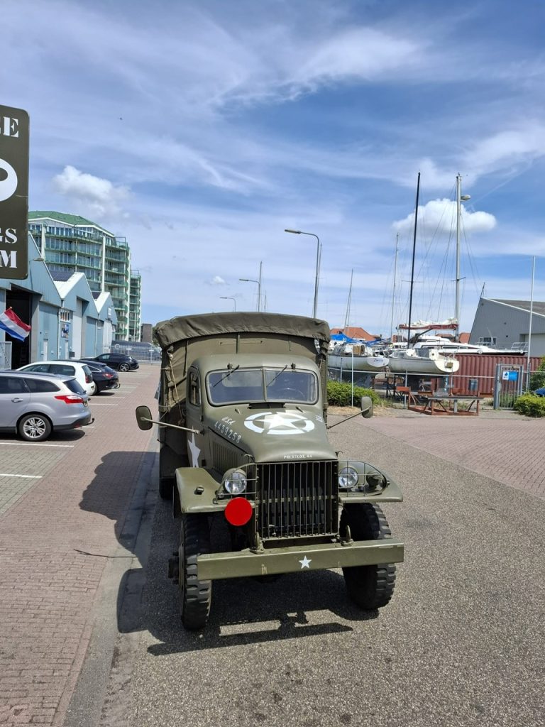 Foto van een Amerikaanse legergroene legertruck uit WO II, die geparkeerd staat langs een straat in een havengebied. Het voertuig heeft een witte ster en identificatienummer op de motorkap en voorbumper. Het heeft een canvas overkapping aan de achterzijde en een kenmerkende gietijzeren grille vooraan. Achter de truck zijn moderne auto's en een gebouw te zien, met verderop in de achtergrond zeilboten en jachthavenstructuren onder een deels bewolkte, blauwe lucht.