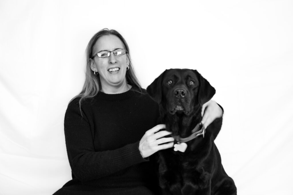 Zwart-wit foto van Debby en haar blindengeleidehond Tarka. Debby zit op een bankje voor een witte achtergrond. Ze heeft lang haar en draagt een zwarte trui en een bril. Ze heeft haar armen om zwarte labrador Tarka die naast haar zit. Ze kijken beiden recht in de camera.
