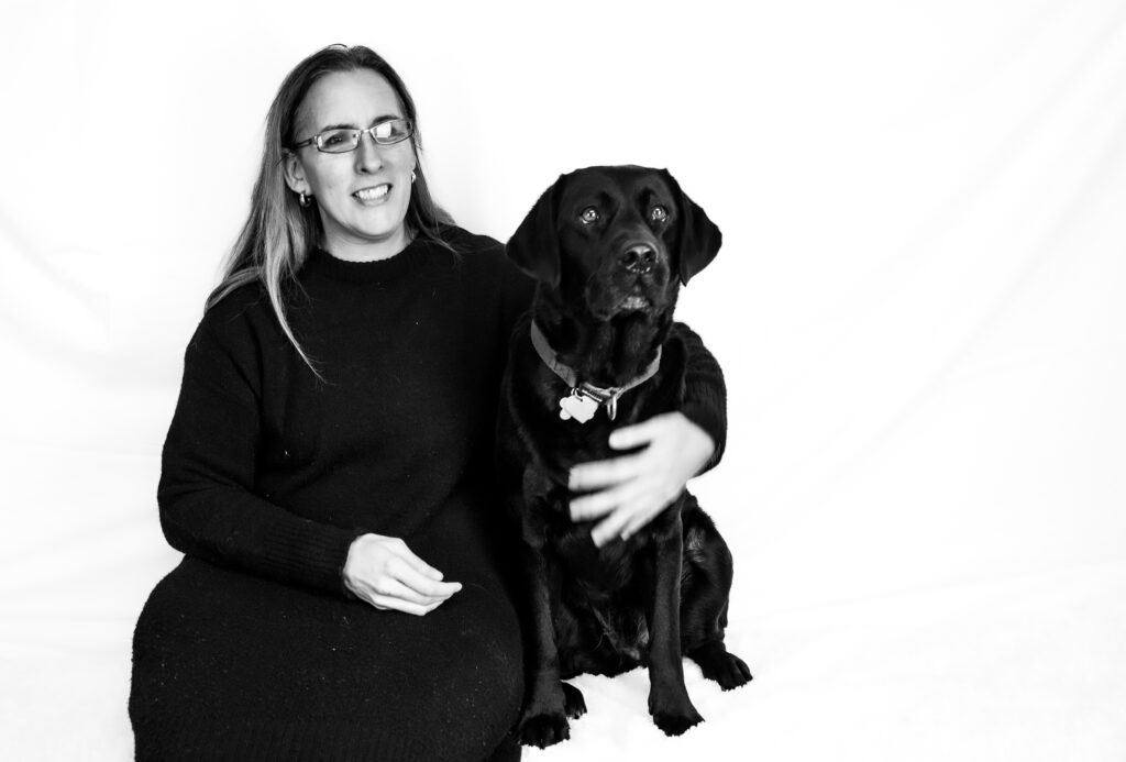 Zwart-wit foto van Debby die naast haar zwarte labrador Tarka zit. Debby draagt een bril en heeft lang haar. Ze glimlacht en draagt een donkere trui. Tarka draagt een halsband met een tag. Debby heeft een hand op de schouder van Tarka. Beiden kijken naar de camera. De achtergrond is egaal wit, wat hen duidelijk laat opvallen.
