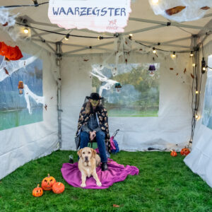 Foto van Debby die groene schmink op haar gezicht heeft en is verkleed als heks. Ze zit, tijdens de Halloween Hondentocht van 2018, in de waarzeggerstent van KNGF Geleidehonden met blonde labrador Tobias tussen haar benen op een kleed. Er liggen pompoenen om hen heen op de grond.