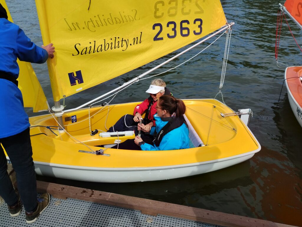 Foto van Debby die samen met een vrouw in een kleine, gele zeilboot zit, die aangemeerd ligt aan een steiger in een haven. De zeilboot heeft een felgeel zeil waarop in grote letters ‘Sailability.nl’ staat en het nummer ‘2363’. Debby draagt een blauwe jas en haar zeilbuddy draagt een rode jas en een witte pet. Ze dragen reddingsvesten. Op de voorgrond staat een derde persoon, die een blauw jack draagt en de boot vasthoudt. Aan de rechterkant zie je nog een deel van een andere zeilboot met een rood zeil. Het water is kalm.
