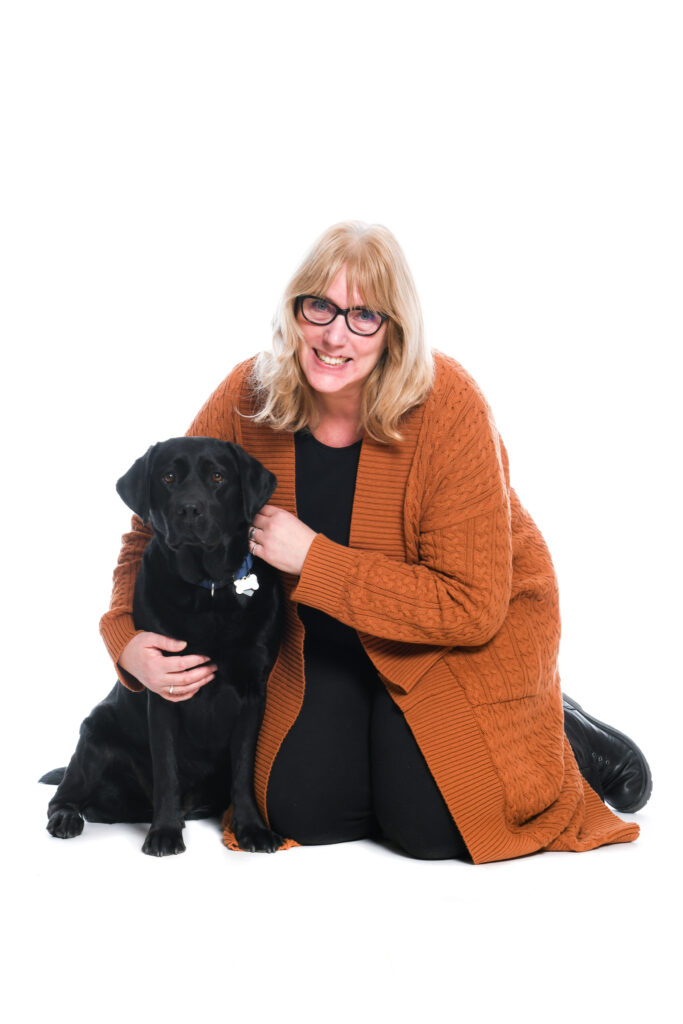 Foto van Debby die op de grond zit met zwarte labrador Tarka. Debby heeft lang haar, draagt een bril en glimlacht naar de camera. Debby heeft haar arm om Tarka heen, die dicht naast haar zit. Ze zitten voor een witte achtergrond.