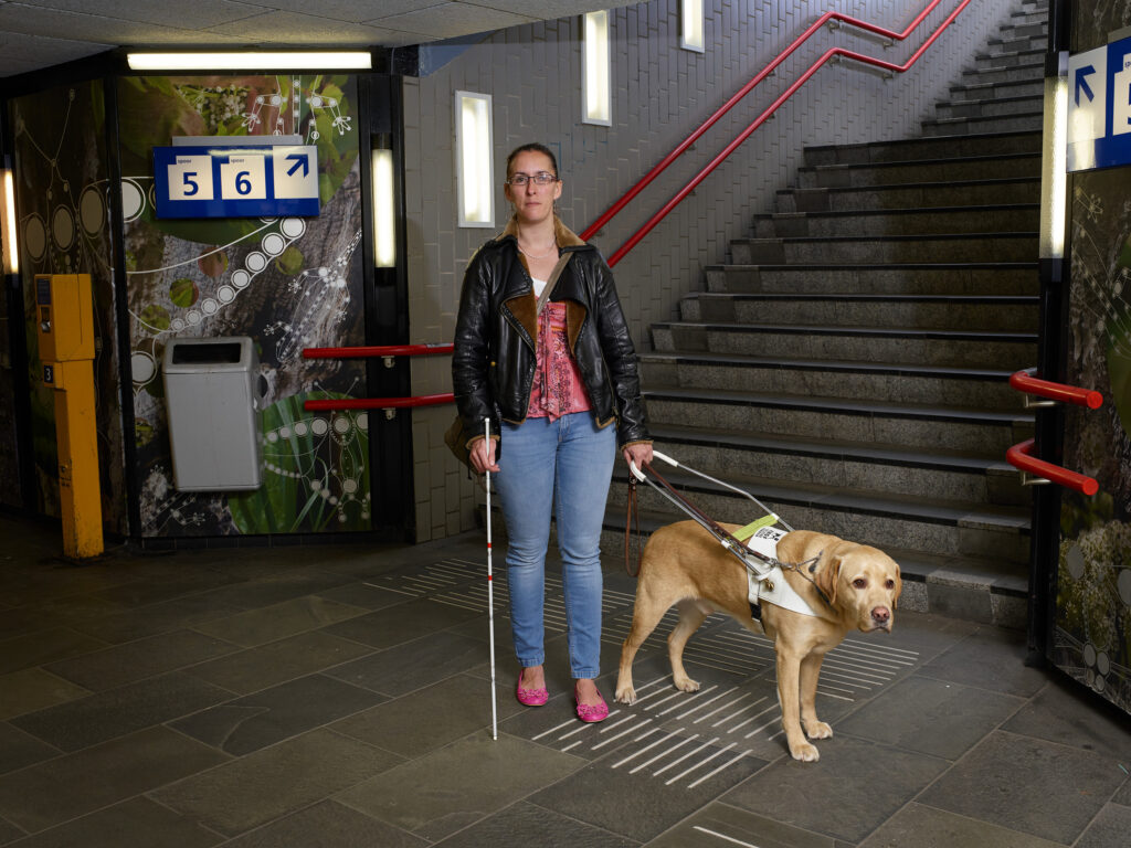 Foto van Debby die samen met blindengeleidehond Tobias onderaan een stationstrap staat. Tobias is een goudblonde labrador reu.