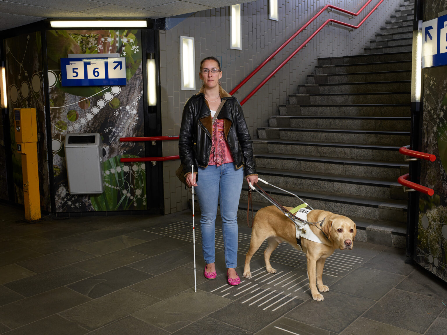 Foto van Debby die samen met blindengeleidehond Tobias onderaan een stationstrap staat. Tobias is een goudblonde labrador reu.