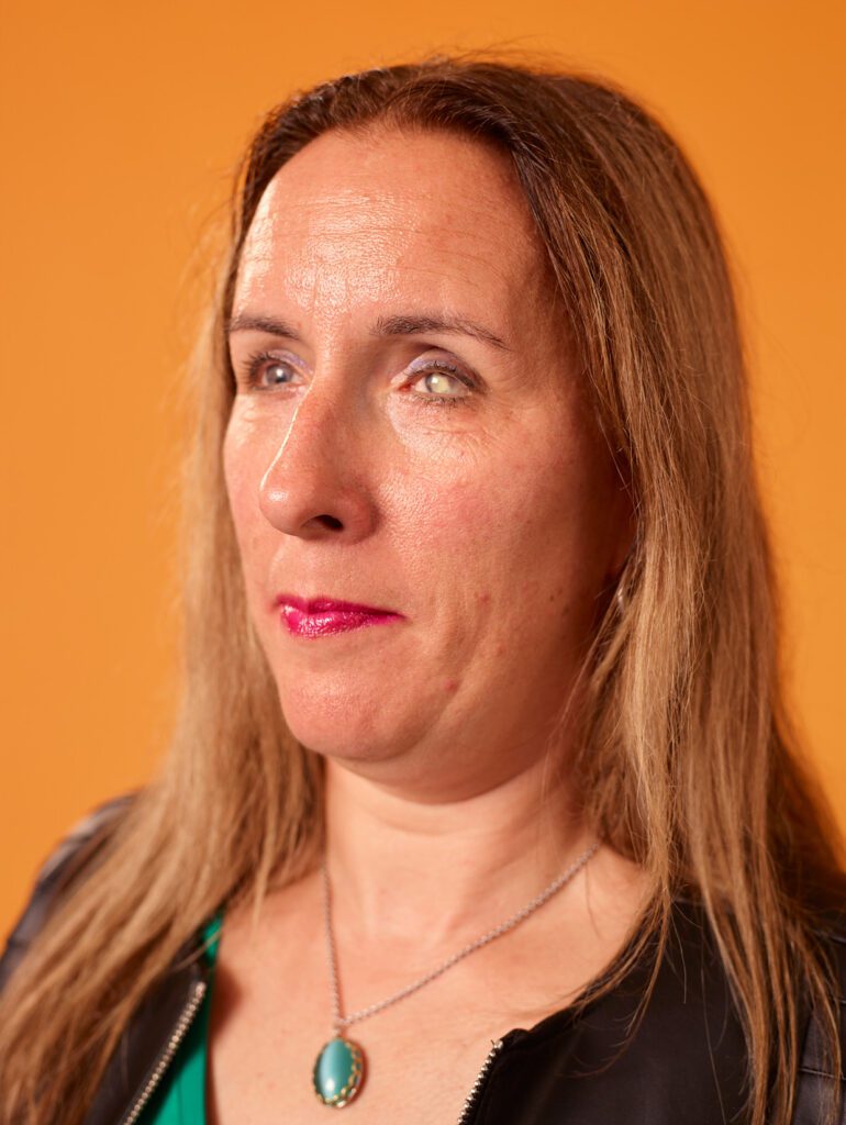 Foto van Debby tegen een oranje achtergrond. Je ziet haar gezicht en een deel van haar bovenlijf. Ze heeft lang, lichtbruin haar en draagt opvallende felroze lippenstift. Ze heeft een zijwaartse blik en een bedachtzame uitdrukking op haar gezicht. Ze draagt een zwart, leren jasje, met een groen shirt eronder. Om haar nek heeft ze een ketting met een opvallende ovalen turquoise hanger. De foto is scherp en professioneel belicht, wat zorgt voor een warme gloed op haar gezicht door de oranje achtergrond.