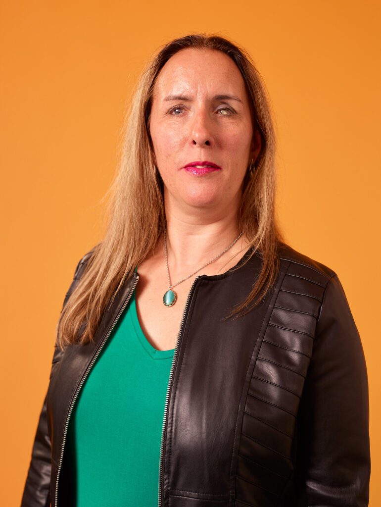Foto van Debby die voor een oranje achtergrond staat. Ze draagt een groene top en een zwart leren jack. Om haar nek hangt een zilveren ketting met een groene hanger en ze draagt felroze lippenstift. De foto is genomen vanaf borsthoogte en ze kijkt direct in de camera met een zelfverzekerde uitdrukking. De belichting is professioneel en gelijkmatig, wat zorgt voor een scherp en helder portret.