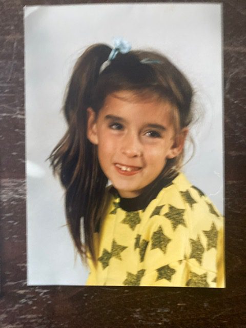 Schoolfoto van Debby als kind. Ze heeft lang, donker haar dat zit vastgebonden in een staart met een lichtblauwe haarband. Ze draagt een geel shirt met zwarte sterren en kijkt naar de zijkant terwijl ze glimlacht. De achtergrond is eenvoudig en wit, wat de focus op Debby legt. Haar uitdrukking heeft een speelse en gelukkige uitstraling.