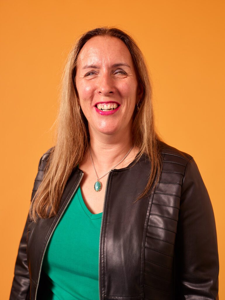 Foto van Debby die lacht. Ze heeft lang, lichtbruin haar en draagt een zwart leren jack met een ritssluiting, over een groen shirt met een V-hals. Ze heeft een zilverkleurige ketting om haar hals met een blauwe steen als hanger en ze heeft felroze lippenstift op. De achtergrond is effen geel.