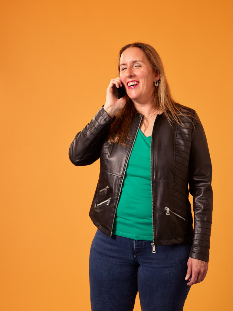 Foto van Debby die een telefoongesprek voert tegen een oranje achtergrond. Ze lacht hartelijk tijdens het gesprek en draagt casual kleding: een zwart leren jack met ritsen, een heldergroen shirt eronder, en een donkerblauwe spijkerbroek. Ze heeft lang, lichtbruin haar en draagt een ketting. Haar houding is ontspannen en natuurlijk, en ze houdt haar mobiele telefoon tegen haar rechteroor. Je ziet haar vanaf halverwege haar dijen. De foto is gemaakt in een studio-omgeving.