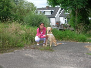 Foto van een hurkende Debby met naast haar blonde labrador Tobias in tuig, die rechtop zit. Op de achtergrond zie je gras en wit-groene Zaanse huizen.