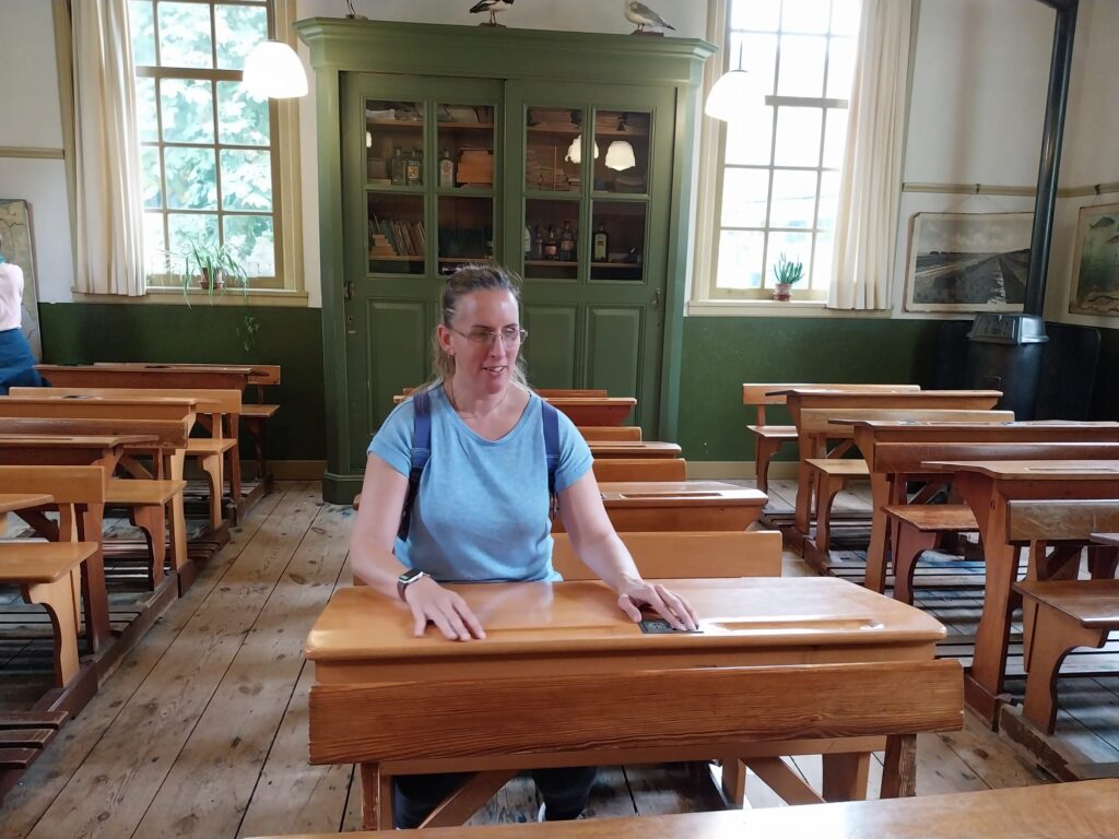 Foto van Debby die in een oud schoollokaal zit. Ze zit op een houten schoolbank aan een houten tafeltje. In het tafeltje zit een gat voor de inktpot en een holte op pennen in te leggen. Debby heeft halflang, bruin haar, draagt een bril en een lichtblauw T-shirt en heeft een paarse rugtas op har rug. . Achter haar zijn rijen van soortgelijke houten schoolbanken te zien. De ruimte heeft houten vloeren en de wanden zijn groen en wit geschilderd. Tegen de achterwand staat een groene kast met glazen deuren, waarin boeken en diverse andere objecten liggen. Erbovenop staan twee vogels. Aan weerszijden van de kast bevinden zich hoge ramen met witte gordijnen die daglicht naar binnen laten. Links naast de kast hangt een schilderij aan de muur. Rechts naast de kast is een zwarte houtkachel te zien, met een groot kunstwerk aan de muur daarboven. De verlichting bestaat uit hangende lampen, één aan elke kant van de kast.