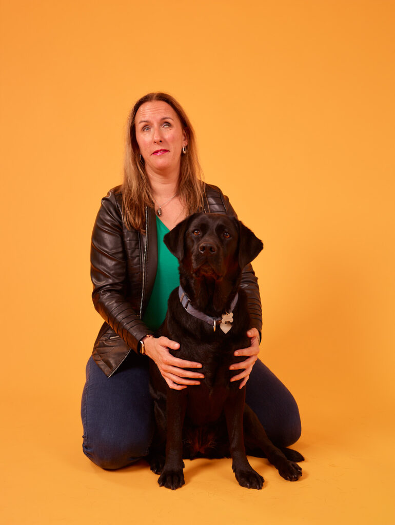 Foto van Debby die op haar knieën zit tegen een effen gele achtergrond. Ze heeft lang, bruin haar en draagt een zwart leren jasje over een groen topje en een blauwe broek. Voor haar zit zwarte labrador Tarka, die ze met beide handen vasthoudt. Tarka kijkt recht in de camera. Debby kijkt omhoog en heeft een rustige uitdrukking op haar gezicht. De achtergrond is effen en zonder enige decoratie.