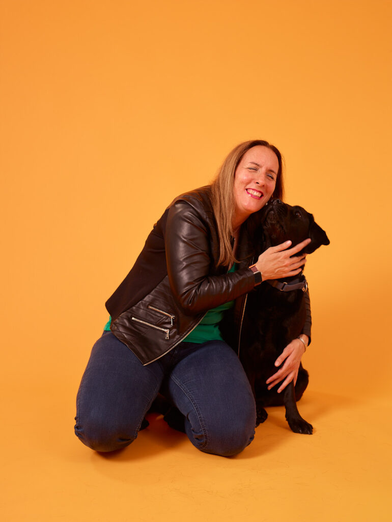 Foto van Debby die op haar knieën zit en haar zwarte labrador, Tarka, knuffelt tegen een felgele achtergrond. Debby heeft lang, lichtbruin haar en draagt een zwart leren jack over een groen shirt, met een blauwe spijkerbroek. Ze lacht breed terwijl ze Tarka stevig vasthoudt. Tarka zit rustig naast Debby en kijkt recht in de camera.