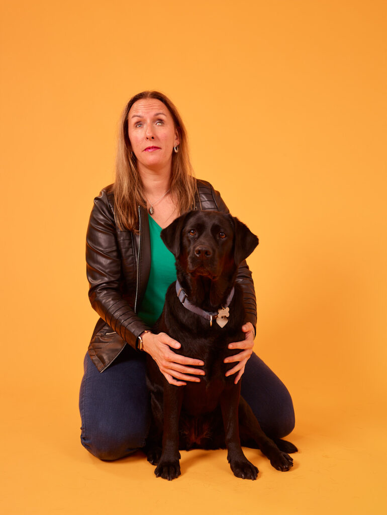 Foto van Debby die op haar knieën zit, terwijl ze zwarte labrador Tarka omarmt. Ze heeft lang, lichtbruin haar en draagt een zwart leren jasje, een groene top en een blauwe spijkerbroek. Ze kijkt enigszins omhoog met een lichte glimlach. Tarka zit voor Debby en kijkt recht in de camera. Debby houdt Tarka stevig vast met beide handen aan de zijkanten van Tarka’s borst. Tarka draagt een halsband met een hartvormige identificatiepenning eraan. Ze zitten voor een gele achtergrond.