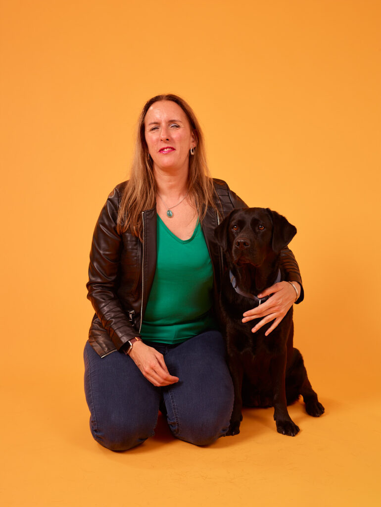 Debby zit geknield op de grond met naast haar zwarte labrador Tarka. Debby heeft lang, bruin haar dat los over haar schouders valt. Ze draagt een groene top, een zwart leren jasje en een donkerblauwe spijkerbroek. Ze heeft een lichte glimlach op haar gezicht en heeft haar arm om Tarka heen. Tarka zit naast haar aan haar rechterkant. Tarka zit ontspannen en kijkt recht vooruit. Beiden kijken schuin richting de camera. De achtergrond is geel.