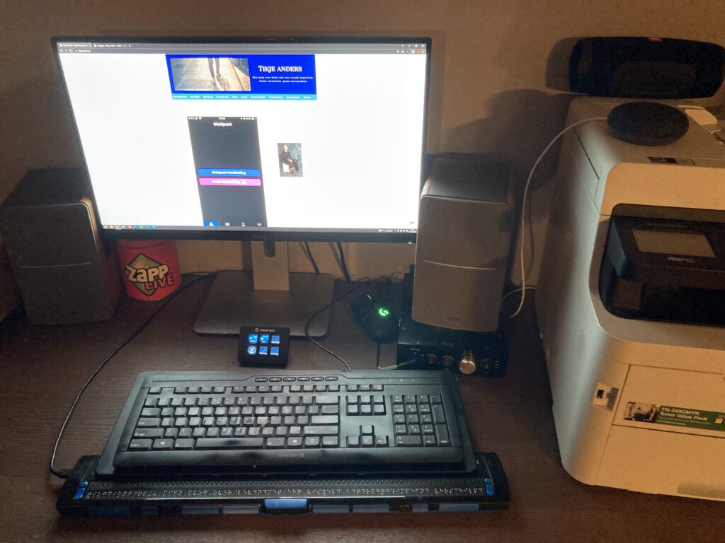 Foto van een bureau met daarop een grote monitor met daarvoor een Stream Deck, toetsenbord en een 80-cellige brailleleesregel.